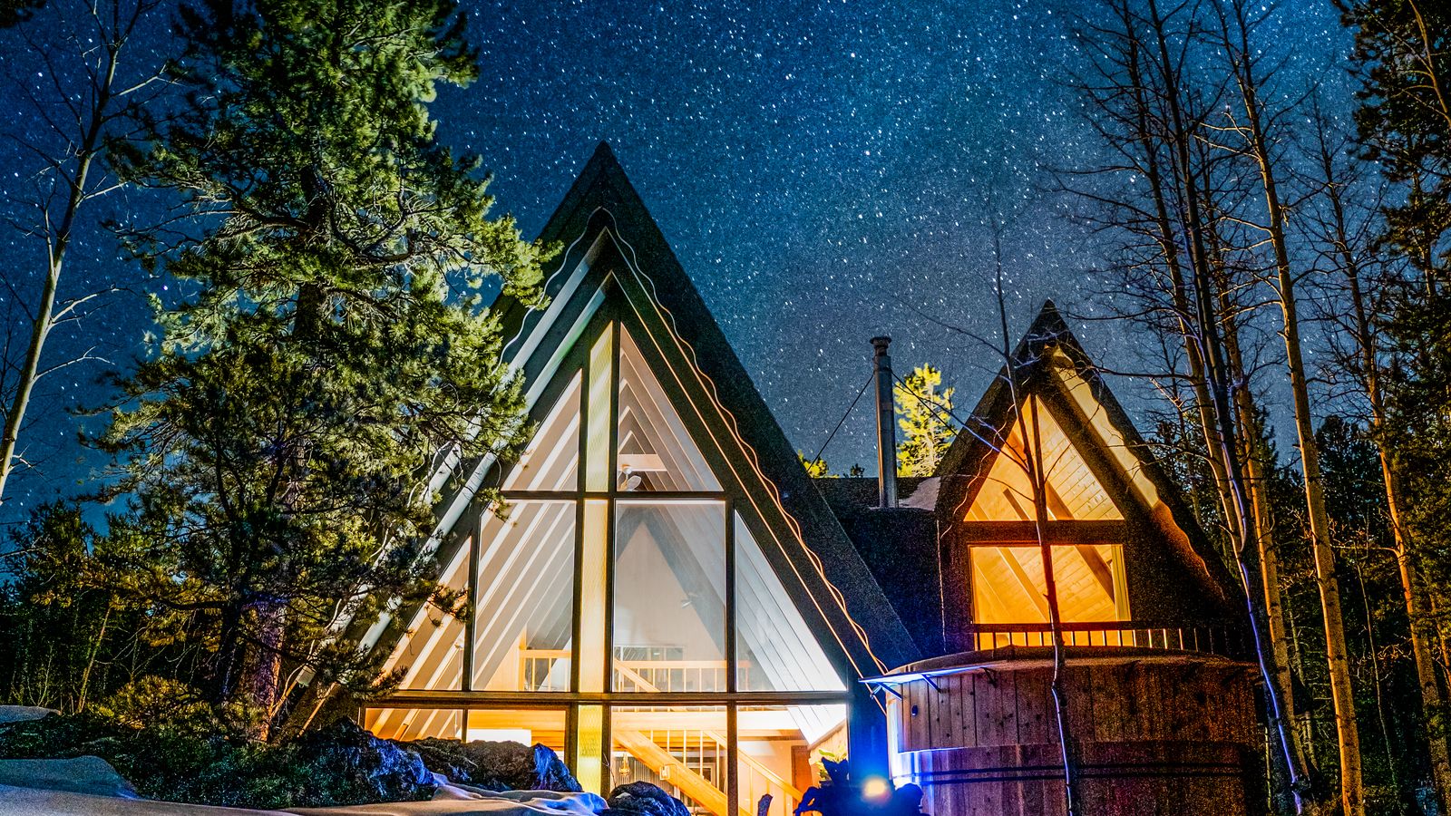 Two lit A-frame cabins in a pine forest under a night sky full of stars