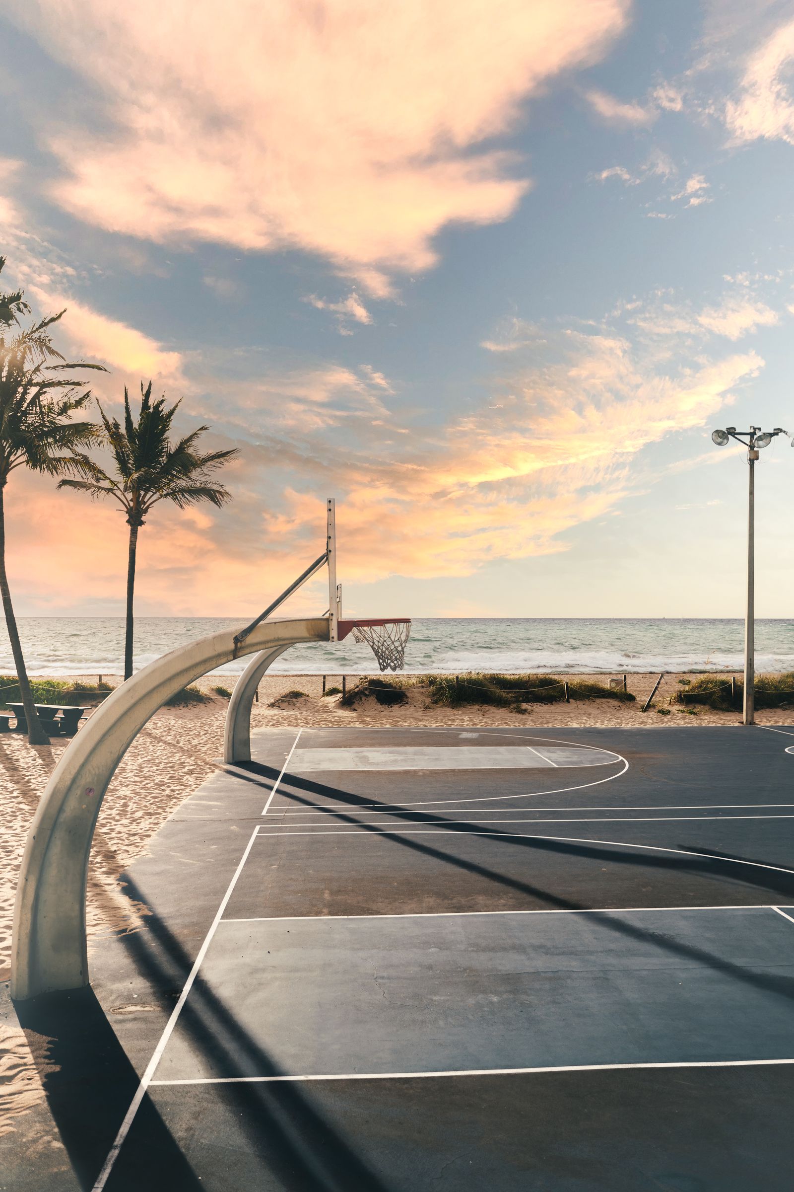 A basketball hoop on a beachside court in Fort Lauderdale, palms and a soft sunset sky behind