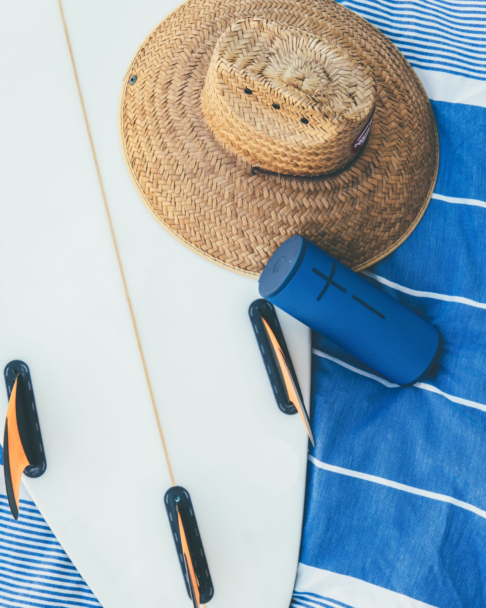 A blue Ultimate Ears speaker on a surfboard with a straw hat and striped towel, from above