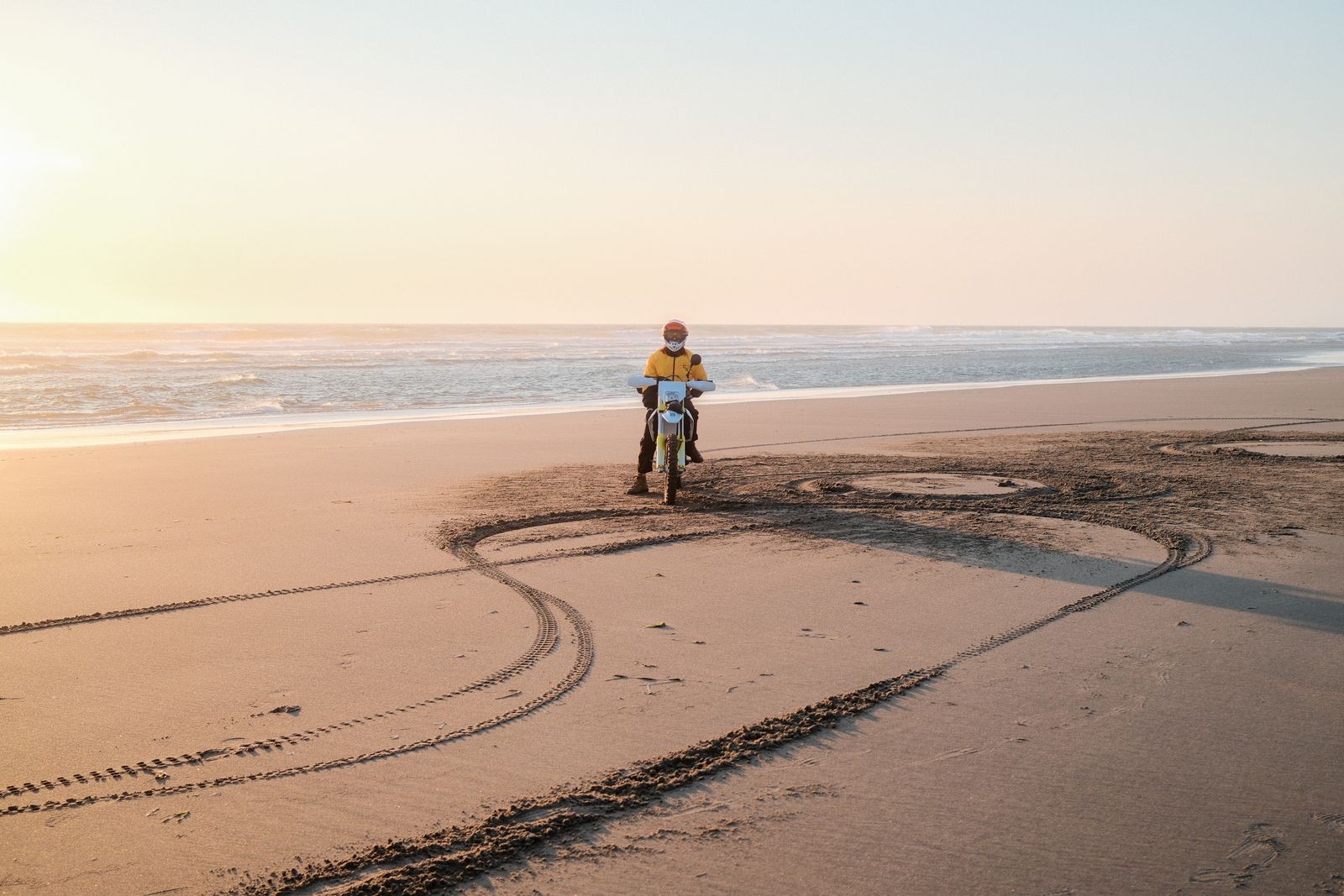 A dirt bike rider at sunset beside looping tire tracks on a wide Oregon beach