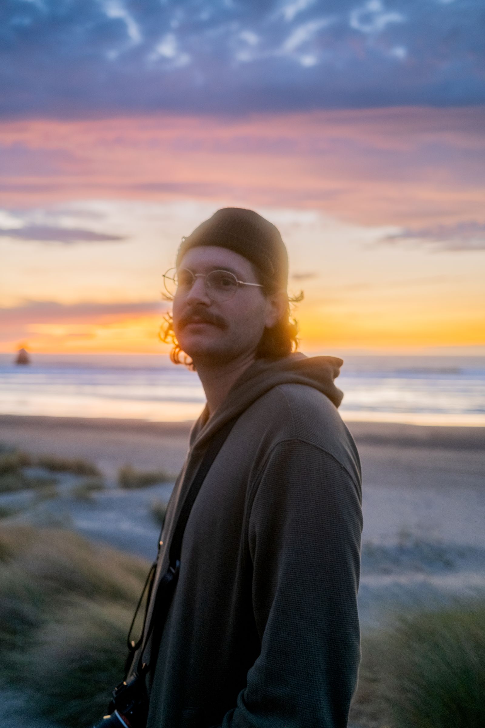 A man in a beanie and glasses on the beach at sunset, pink and gold clouds behind