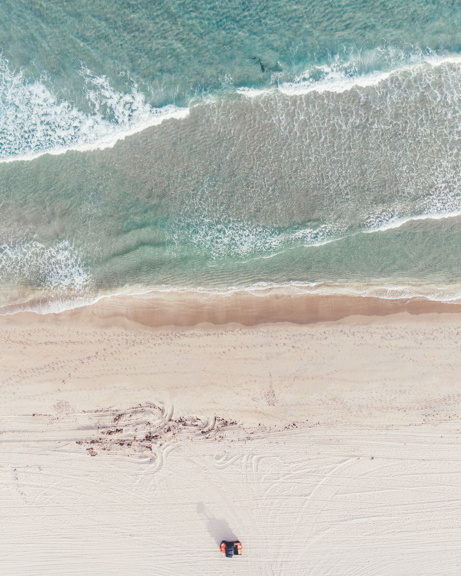Tire tracks on a pale beach beside turquoise surf, from above