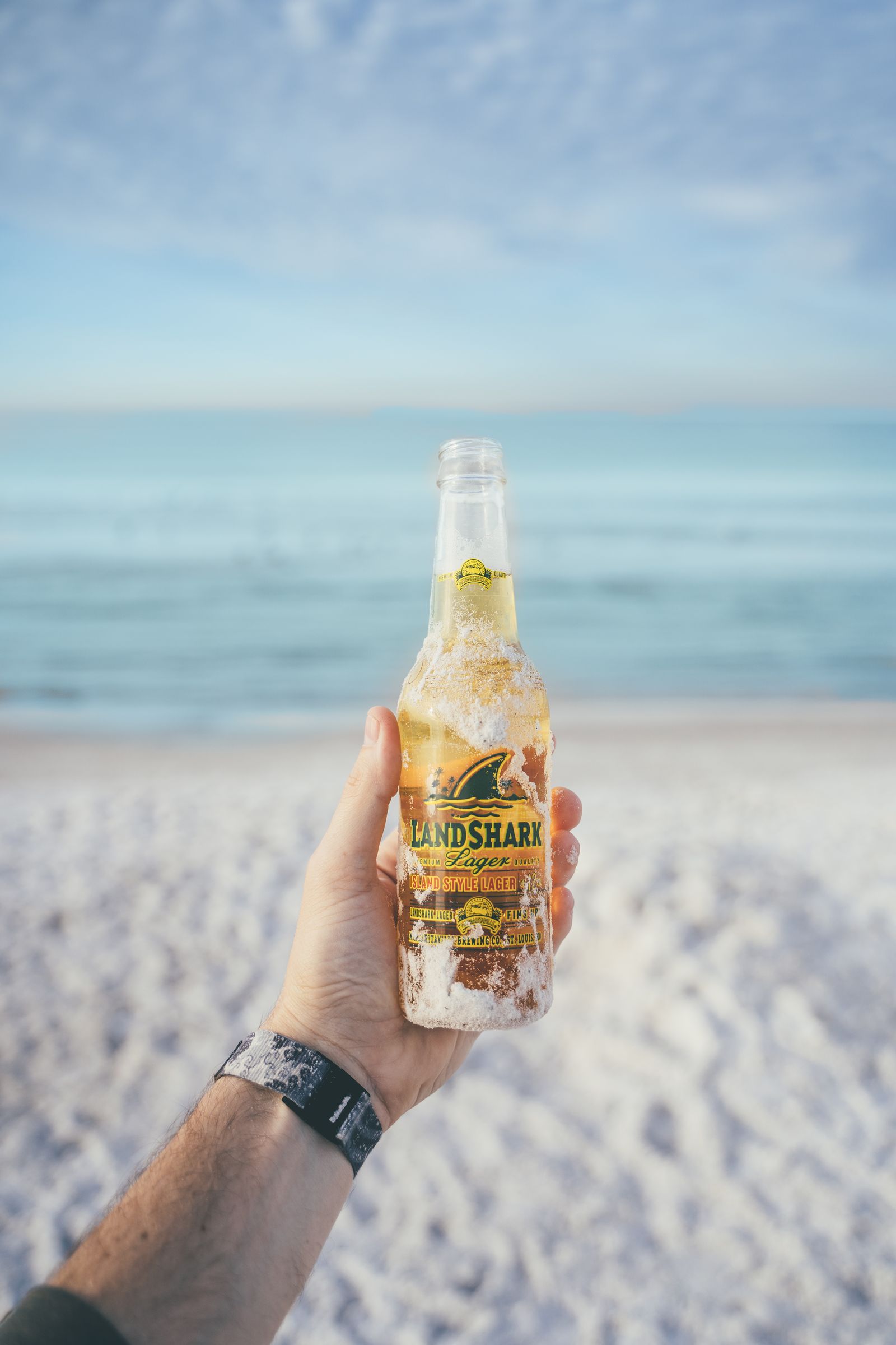 A hand holding up a bottle of Landshark lager over the beach