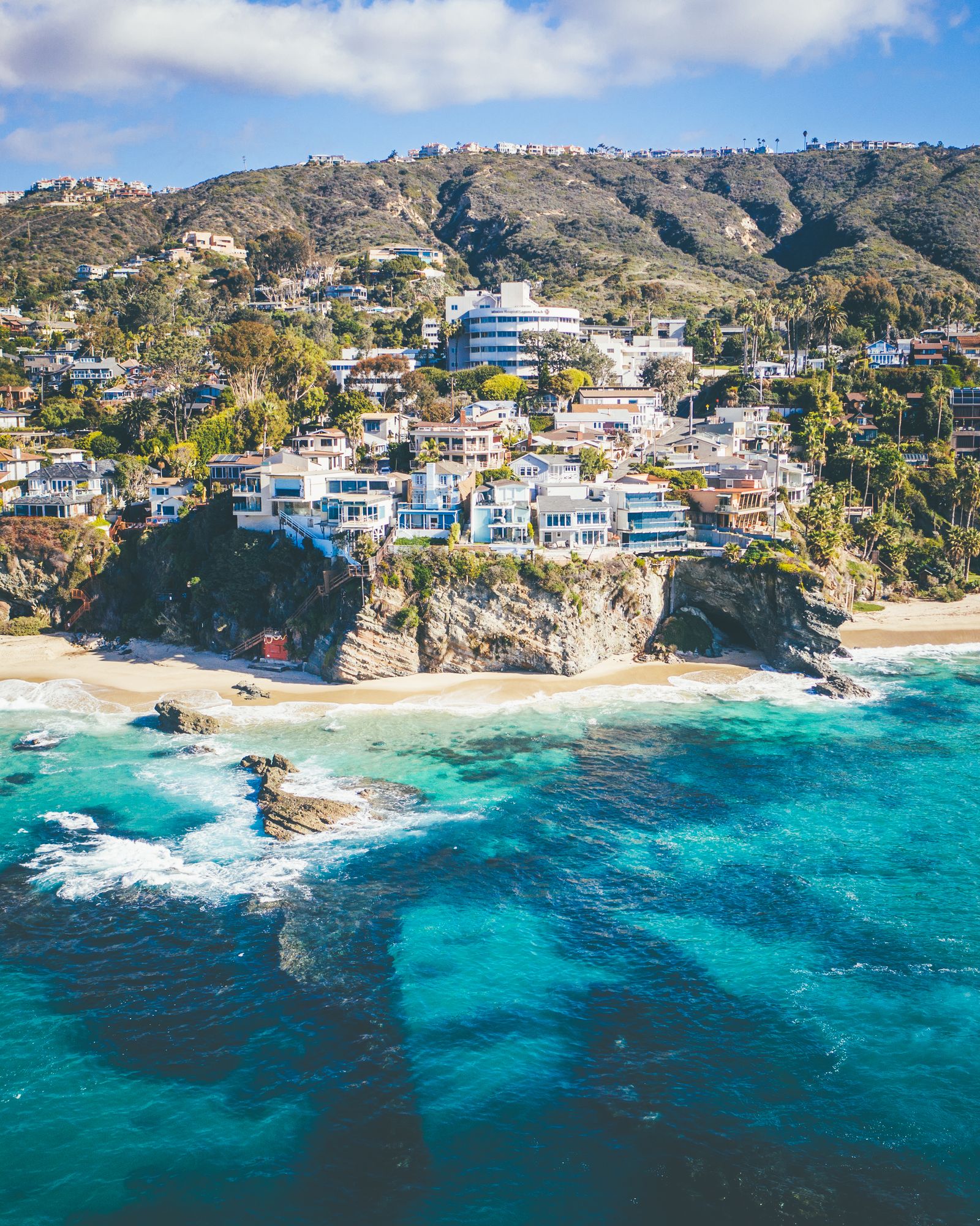 Aerial of a Southern California cove: white houses on a rocky headland above deep blue water and a small sandy beach