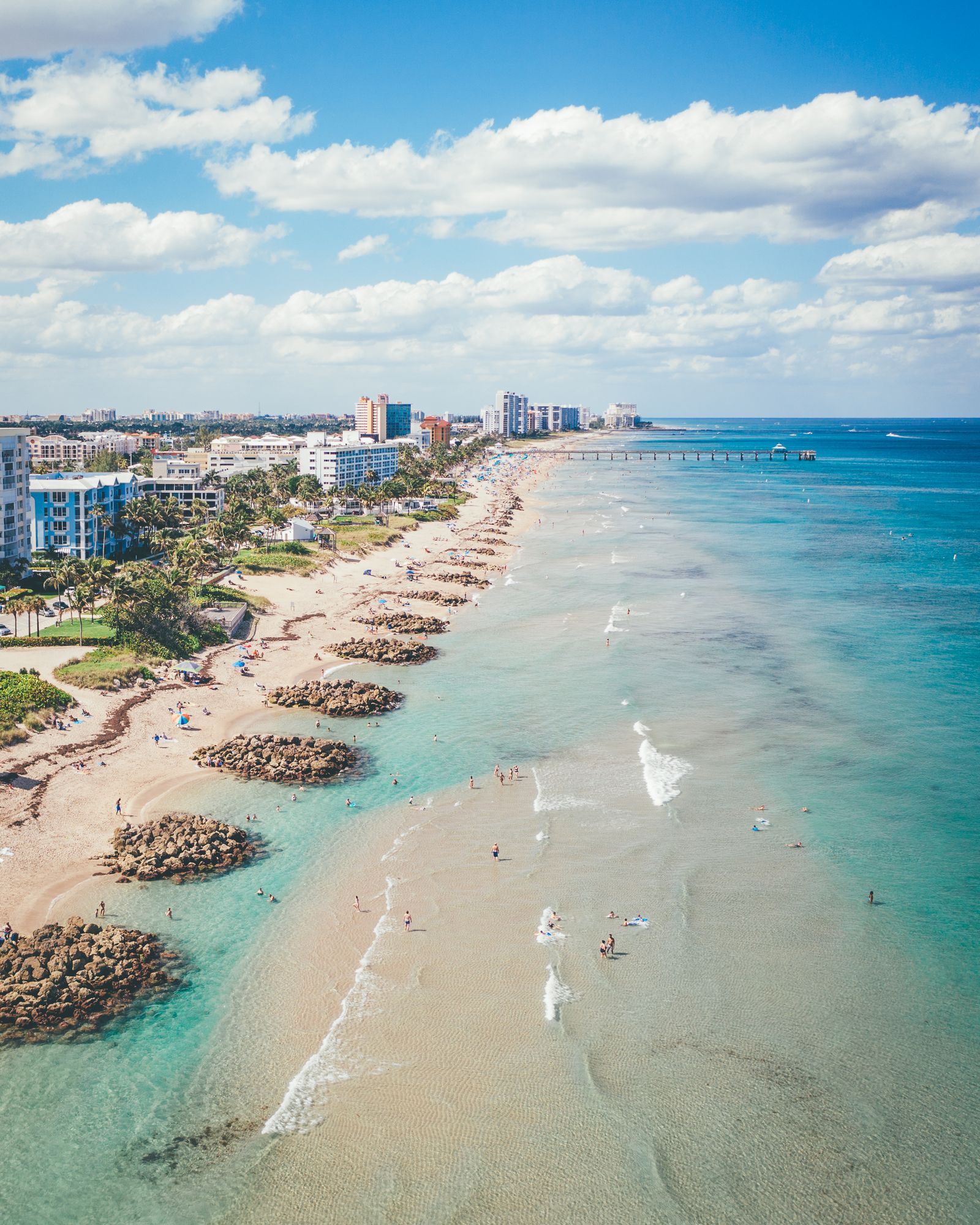 Aerial of Deerfield Beach looking down the shore: rock groins, swimmers, the pier and puffy clouds