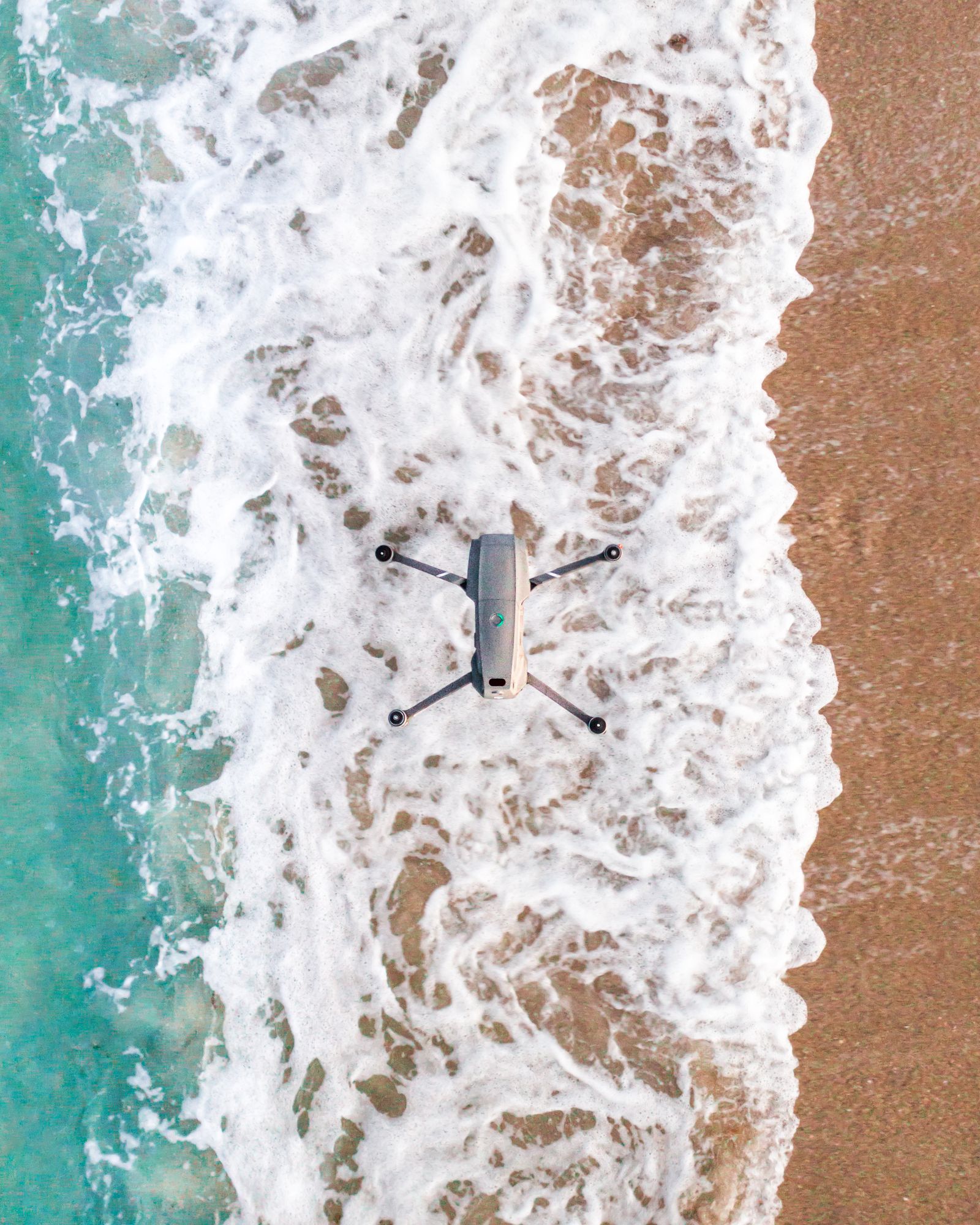 A drone hovering low over white wash and wet sand where a wave rolls up the beach