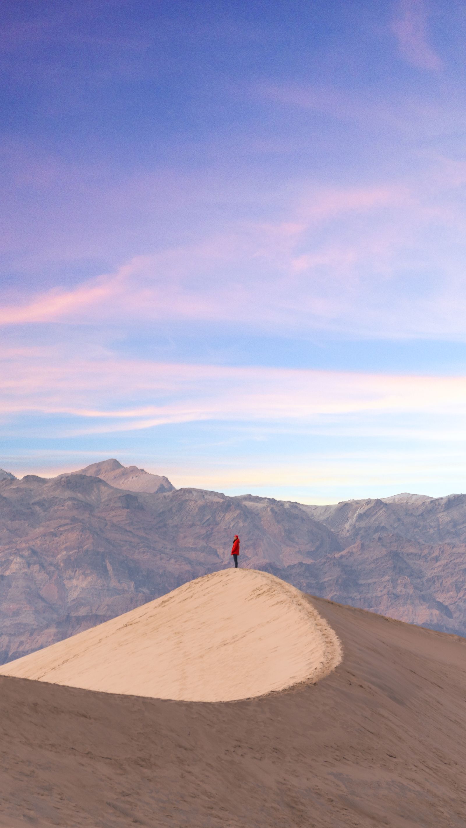 A lone figure standing on top of a tall sand dune at dusk, mountains and pink clouds behind