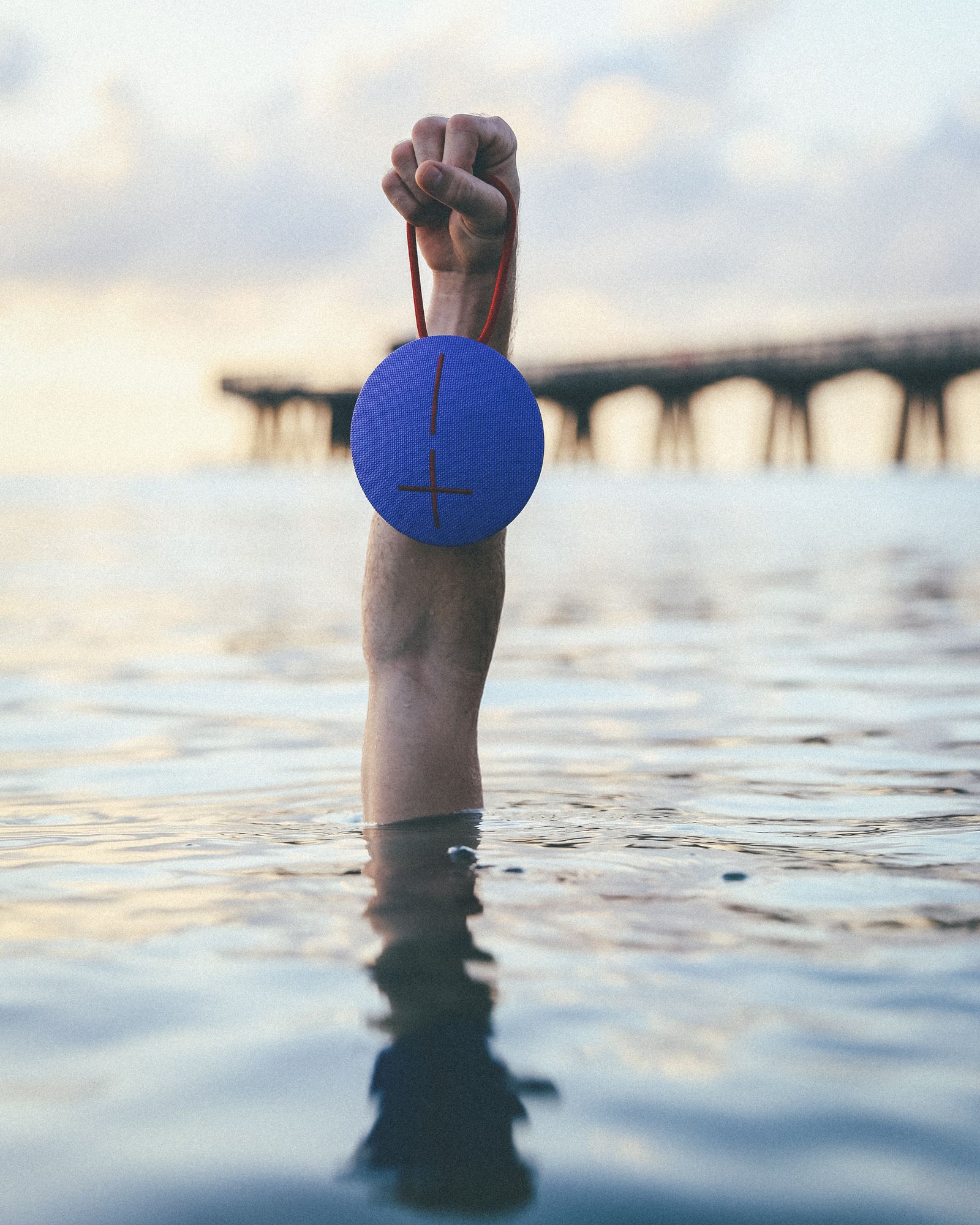 A fist holding a round blue Ultimate Ears speaker by its strap just above the water near a pier