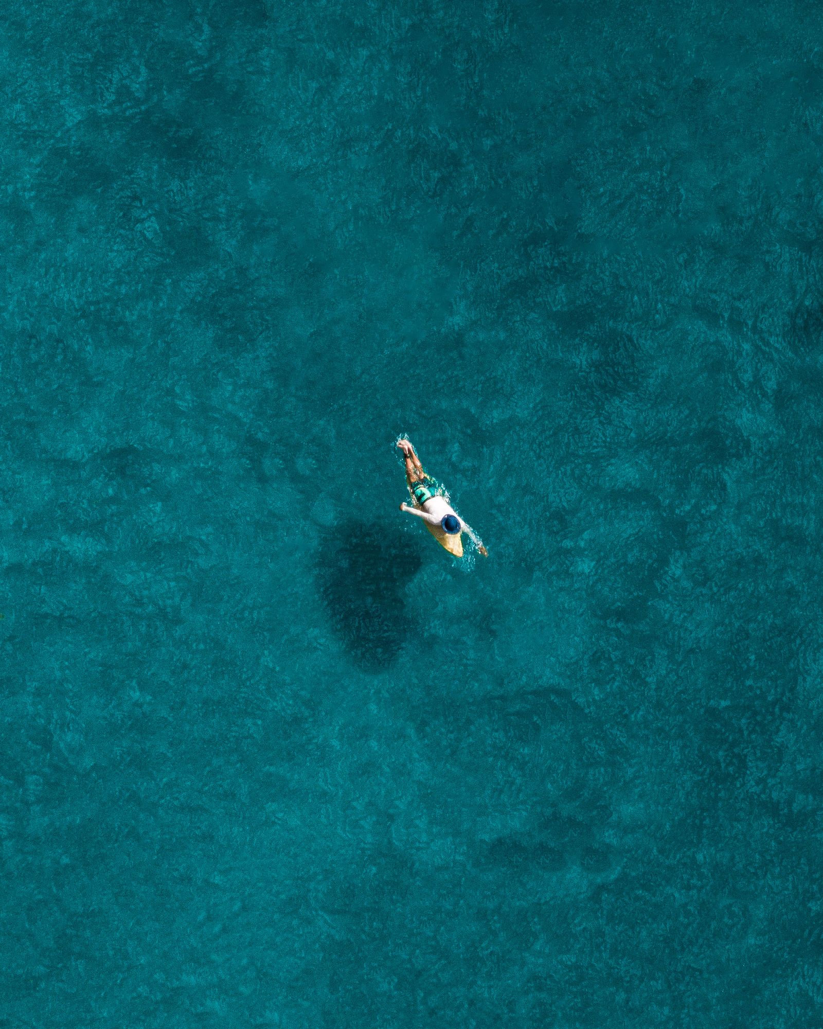 A person floating on a board in deep teal water, seen from straight above