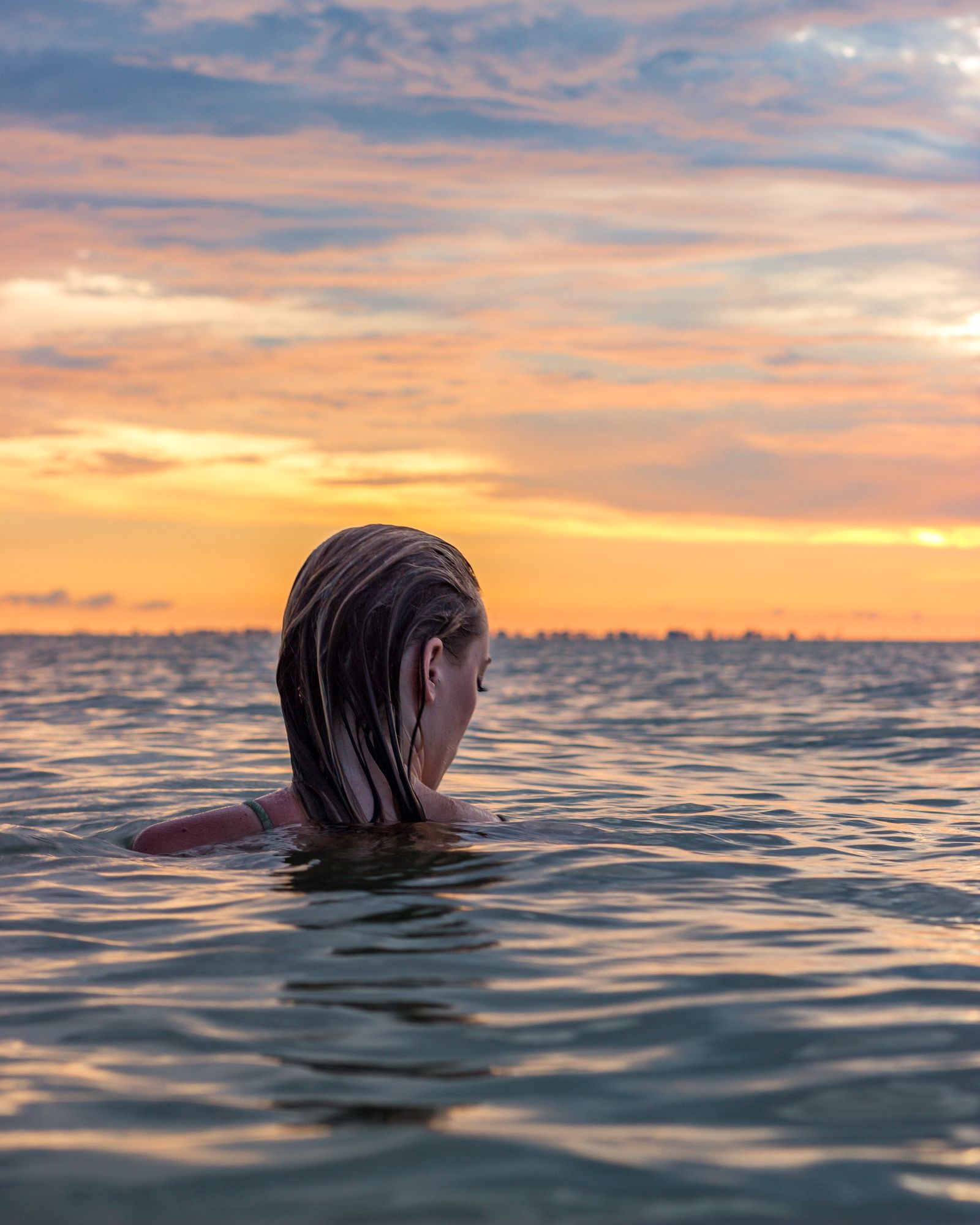 A woman swimming out toward an orange sunset, only her head above the calm water
