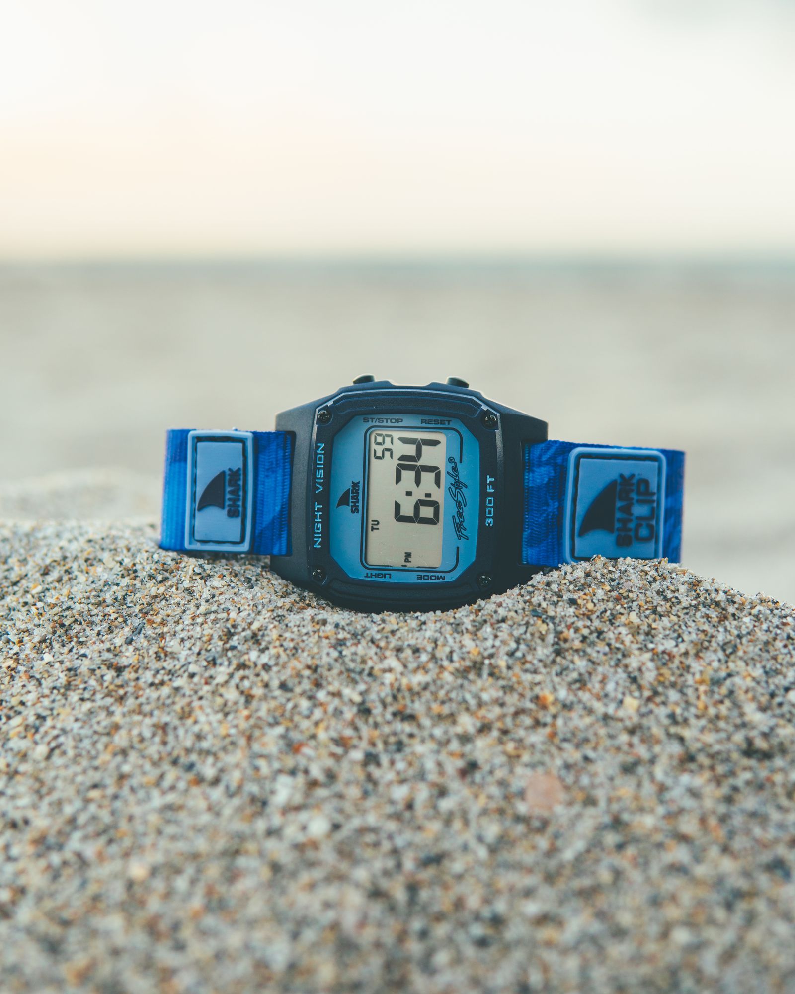 A blue Freestyle watch half buried in wet sand at the shoreline