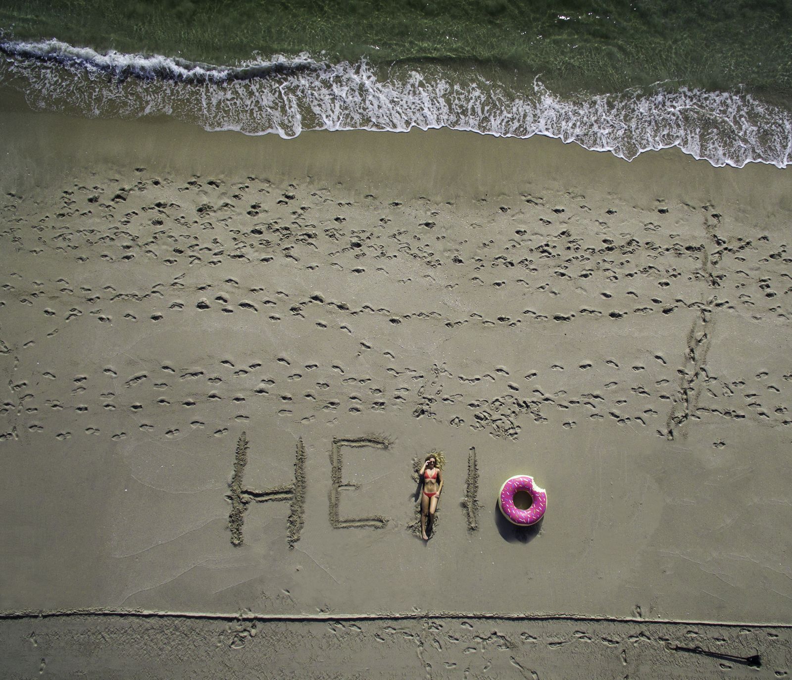 The word HELLO written in wet sand with a pink pool float, from directly above
