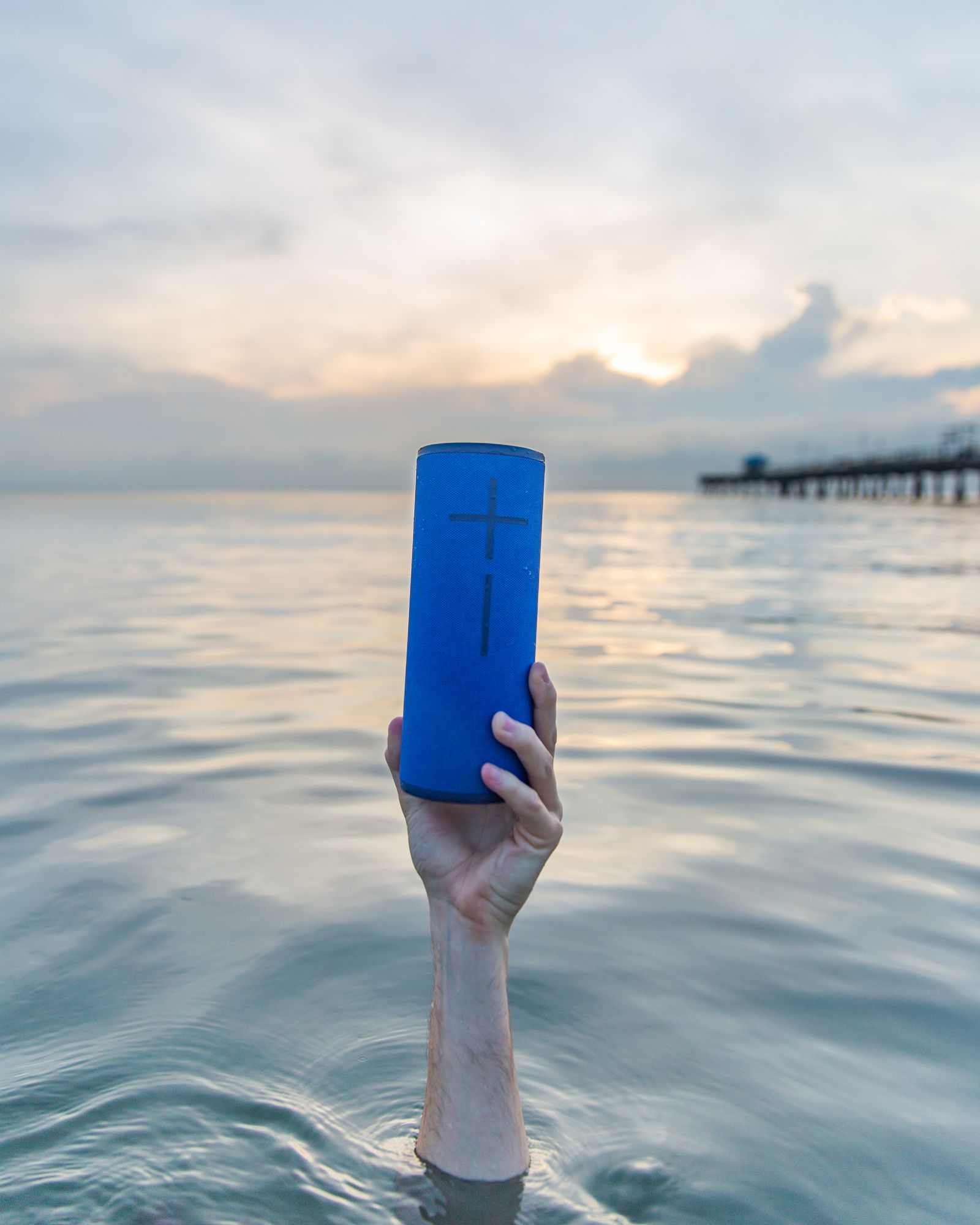 A hand rising out of calm water holding a blue Ultimate Ears speaker at sunset by a pier