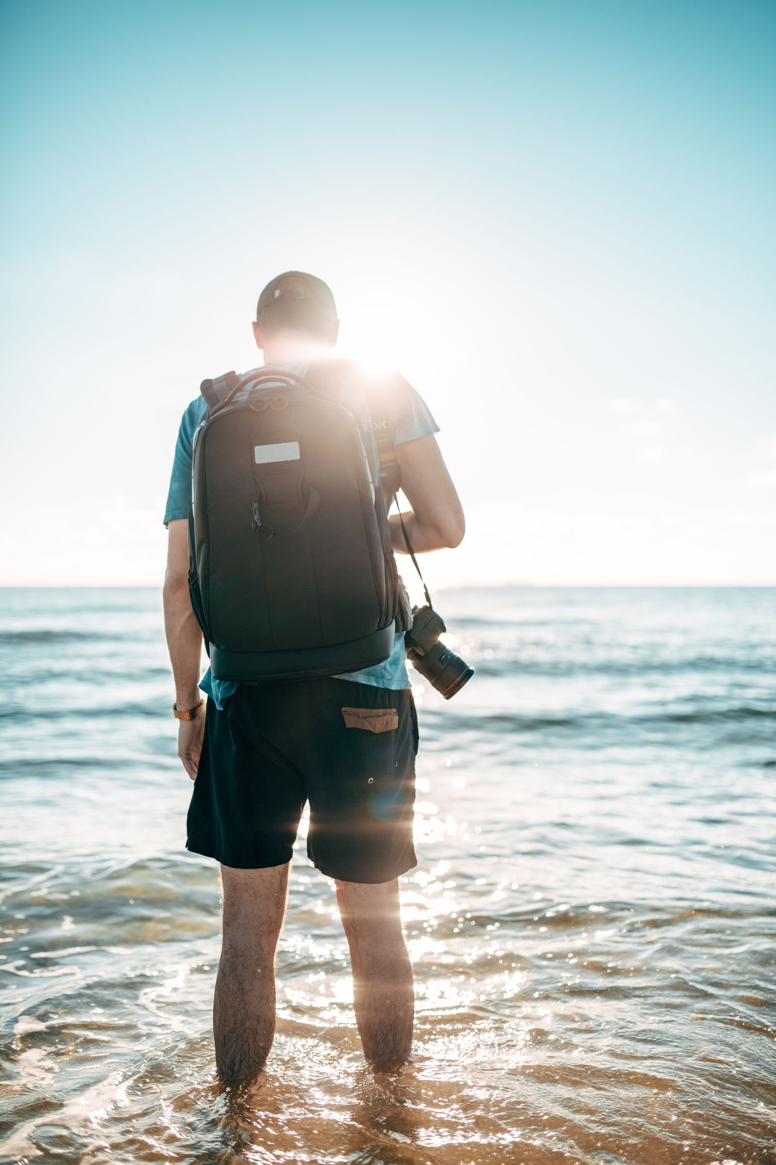 A man with a backpack wading into the ocean toward a bright sun