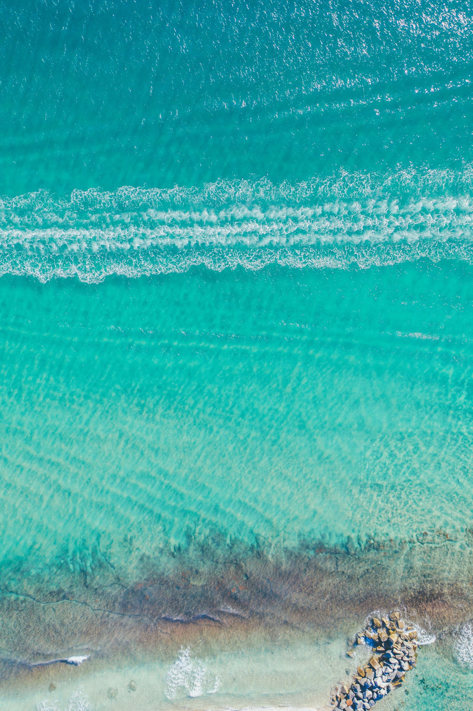 Parallel lines of surf rolling toward a rock jetty in bright turquoise water