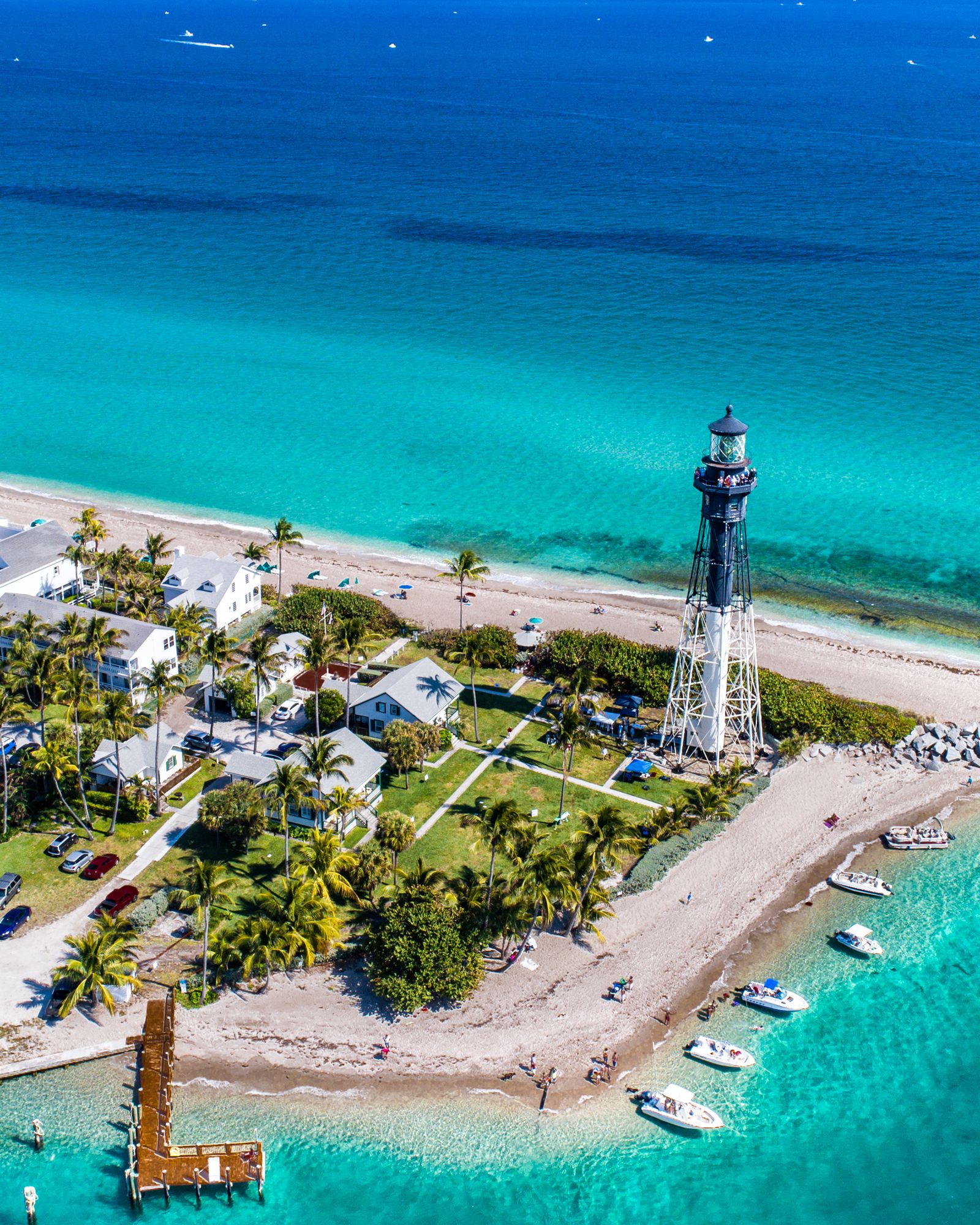 Aerial view of the Hillsboro Inlet lighthouse on a palm-lined point, turquoise water and boats anchored off the beach