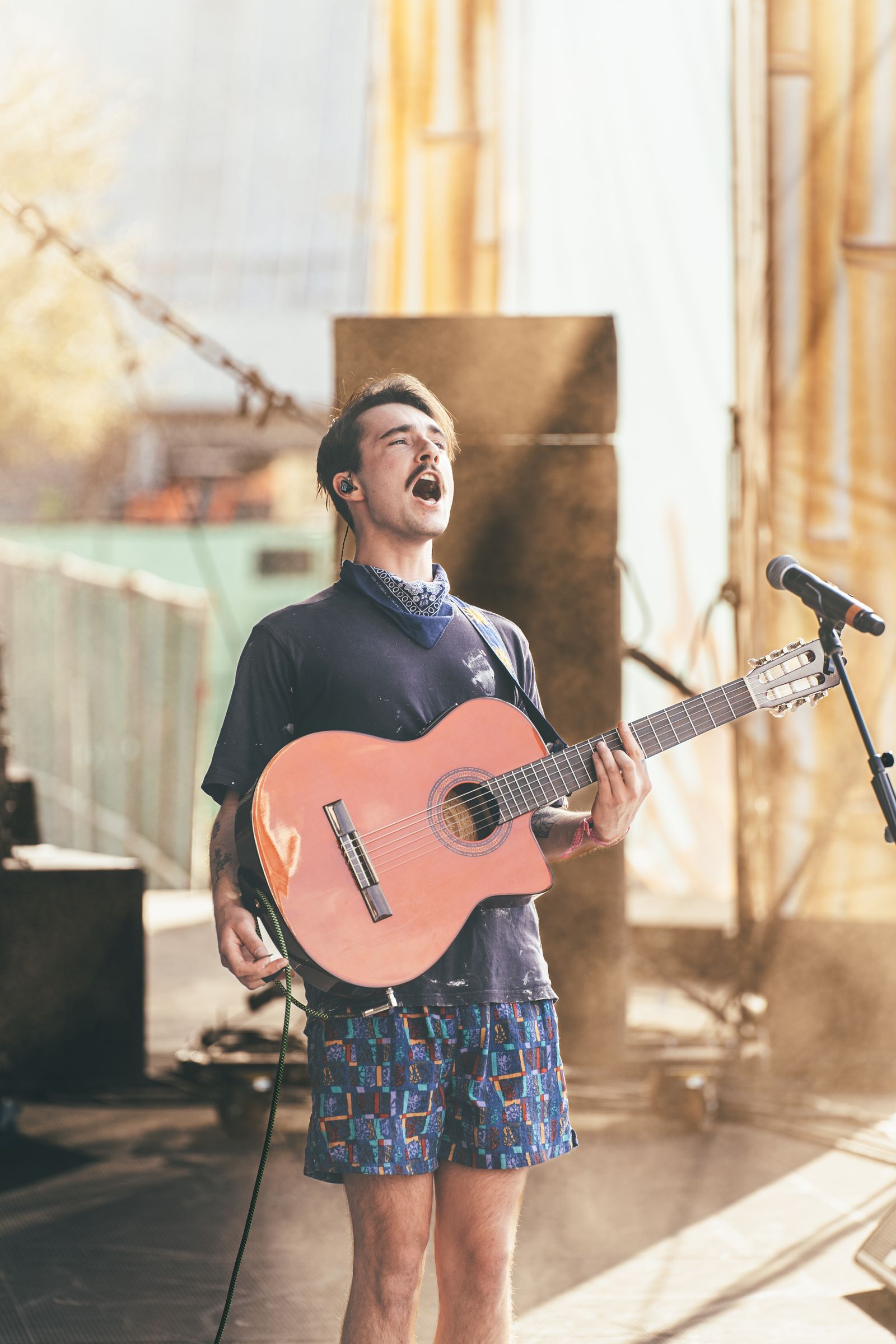 A musician singing with an acoustic guitar on an outdoor stage in soft backlight