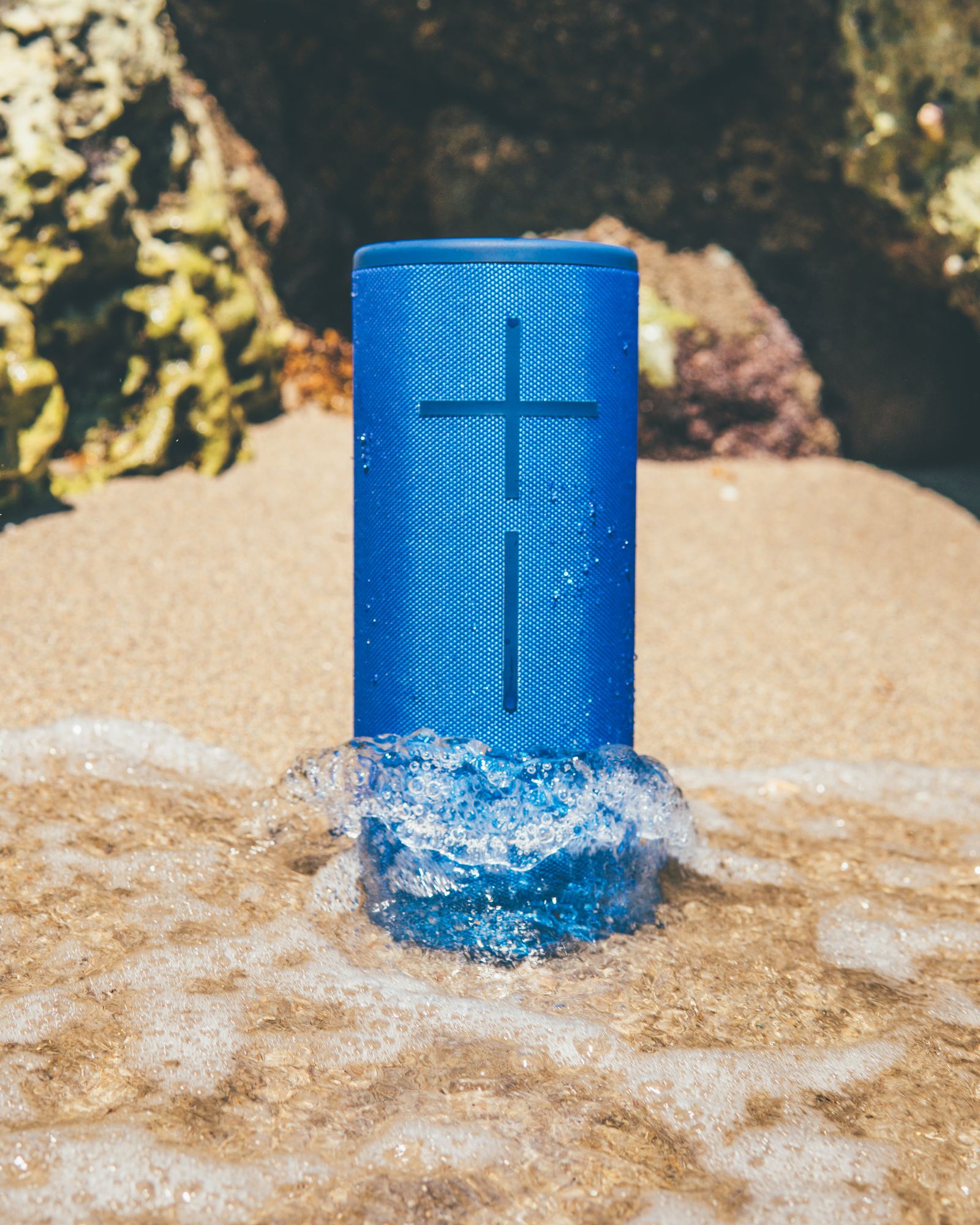 A blue Ultimate Ears speaker standing in the sand as a wave splashes around it