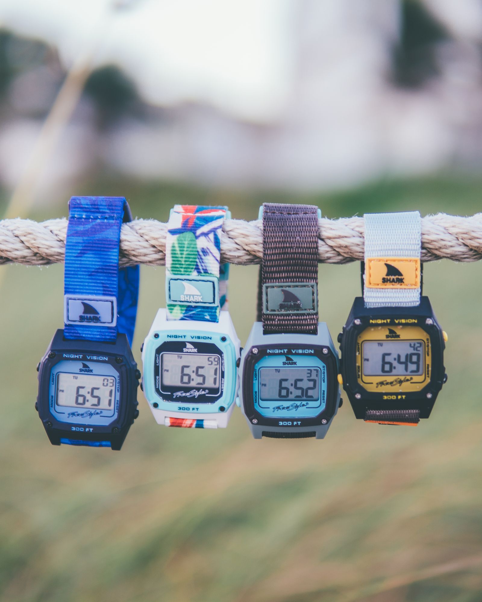 Four Freestyle Shark watches hanging from a rope over a blurred green field