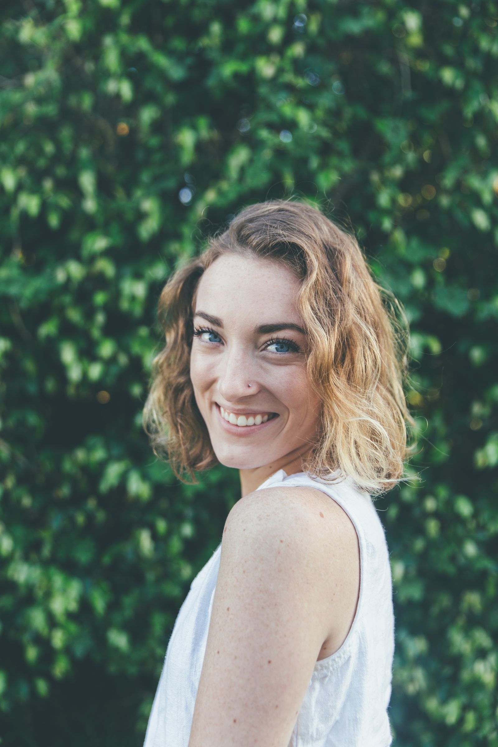 A smiling woman with curly hair looking over her shoulder in front of a green hedge