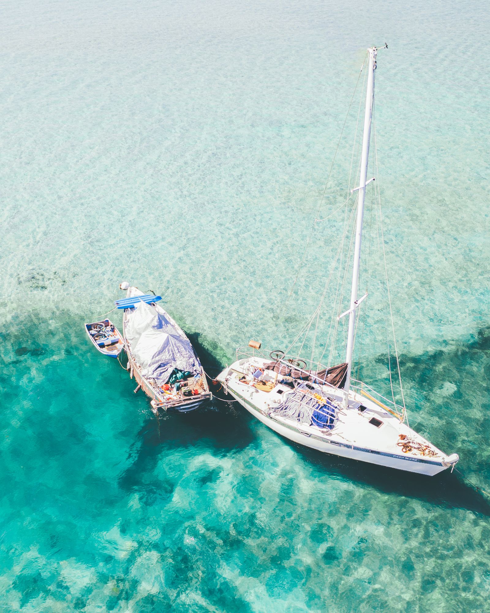 A sailboat and a smaller boat tied together over turquoise water and sand, seen from above