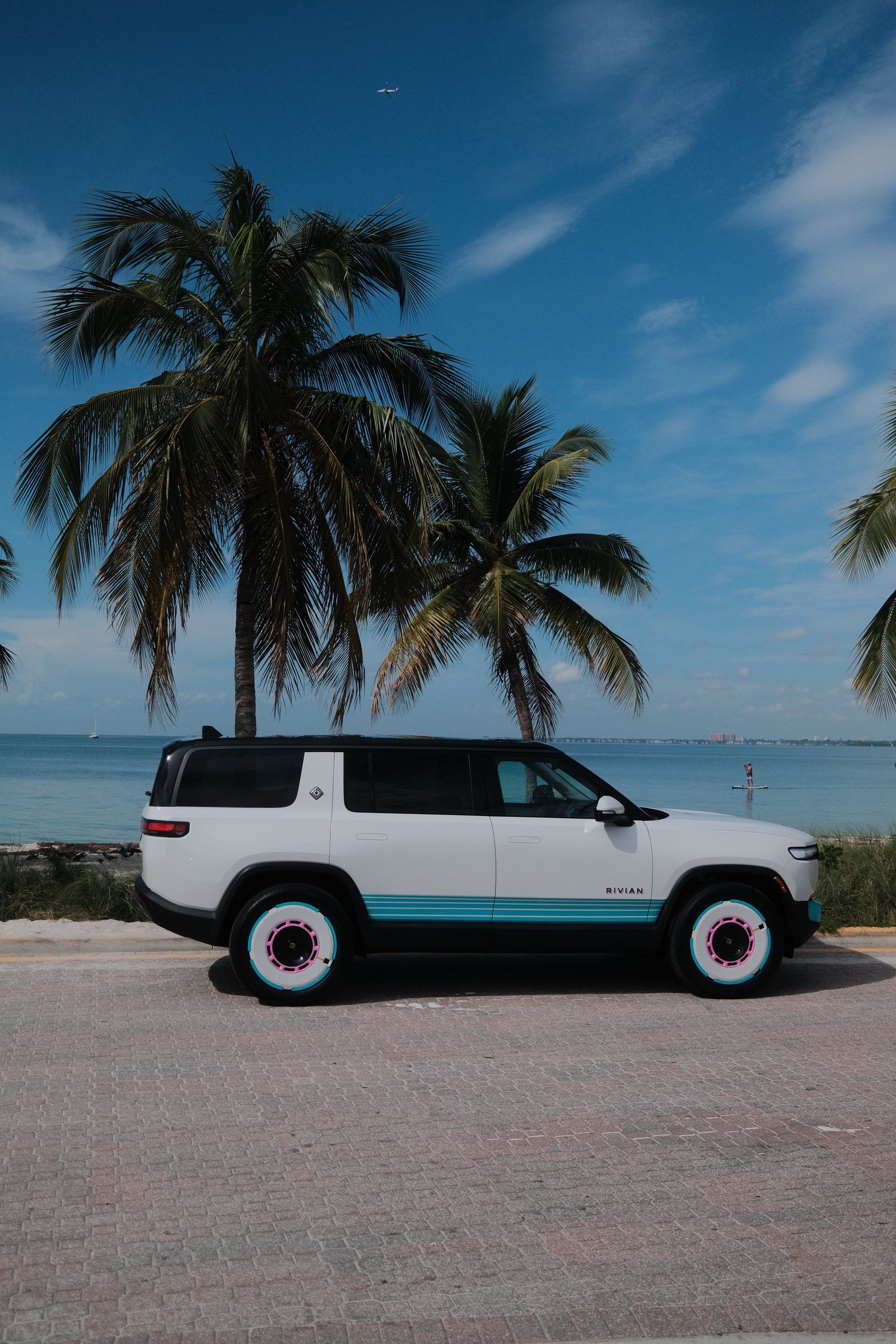 A white Rivian SUV with teal trim parked by the water under tall palm trees