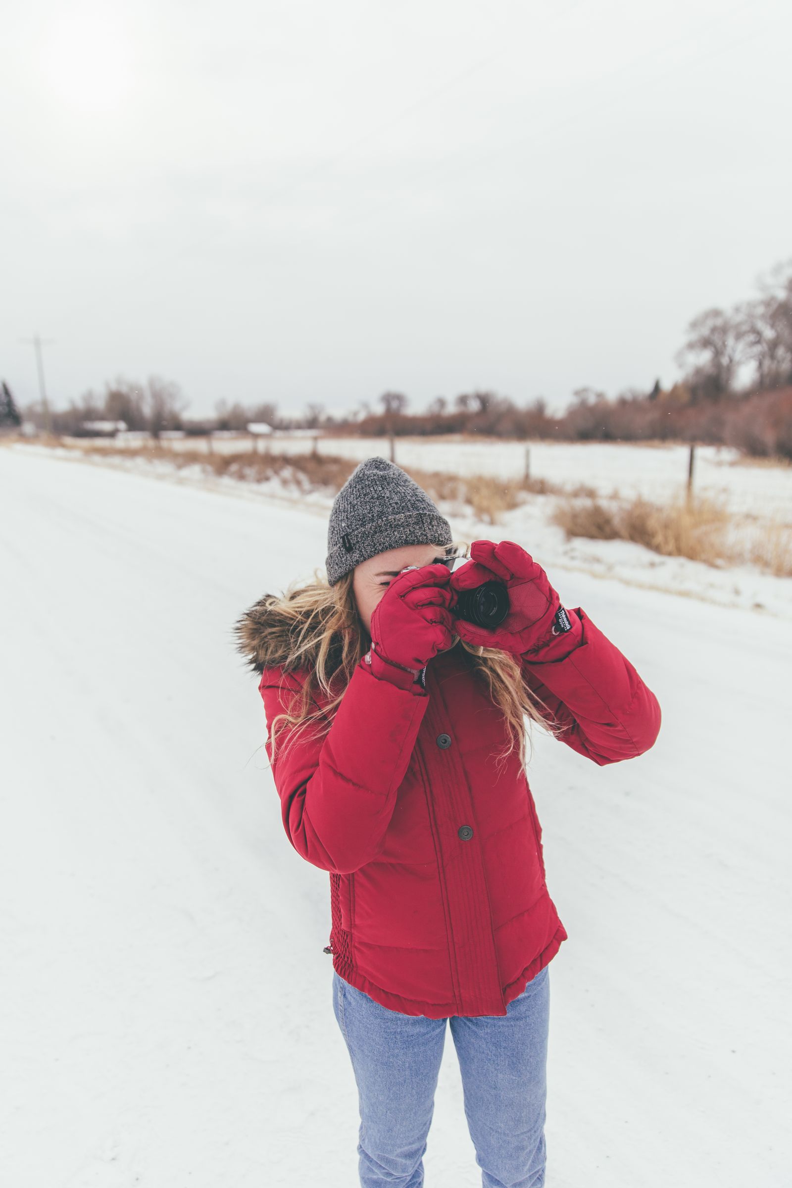 A woman in a red parka and knit hat raising a camera in the snow