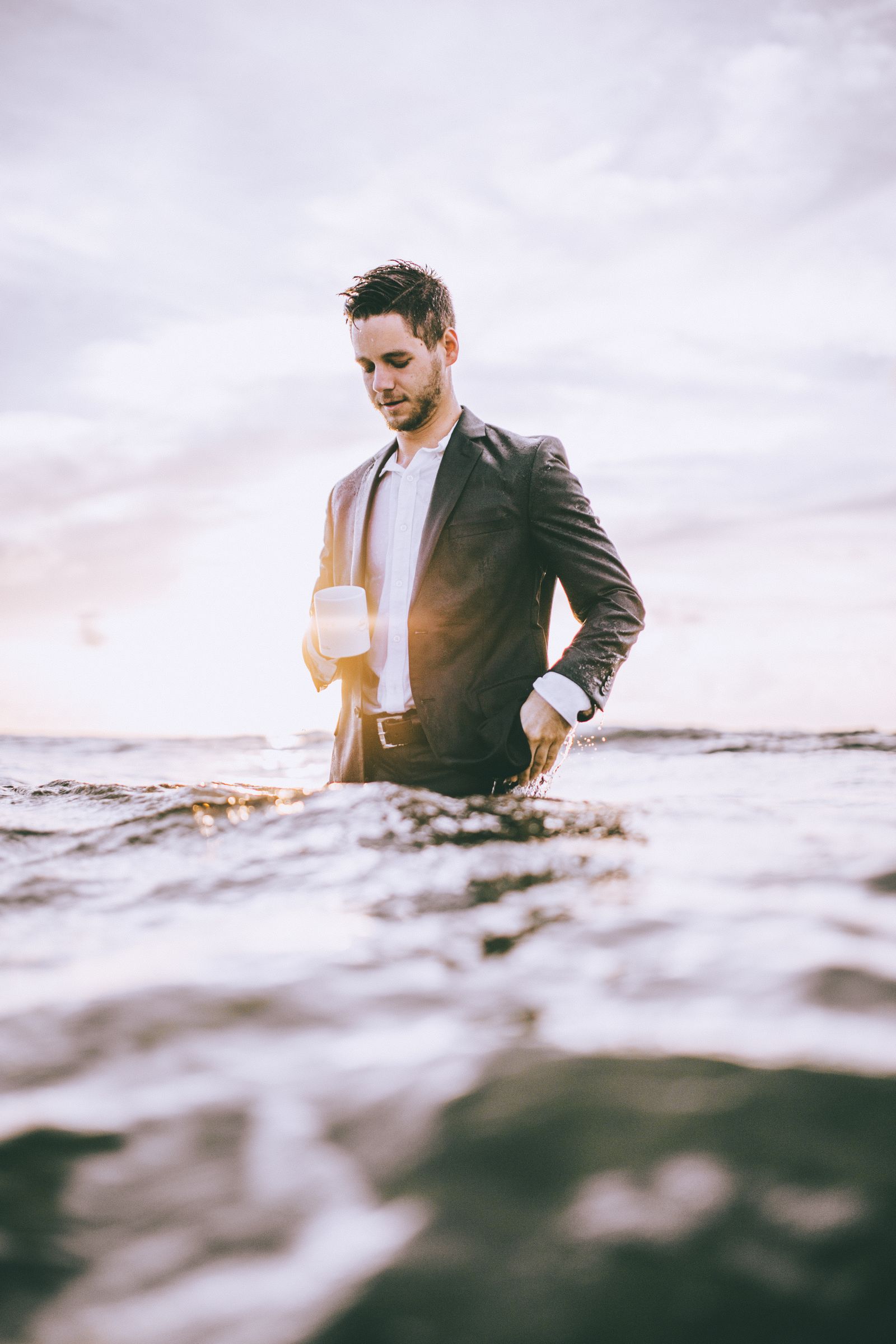 A man in a dark suit standing waist deep in the ocean at sunrise