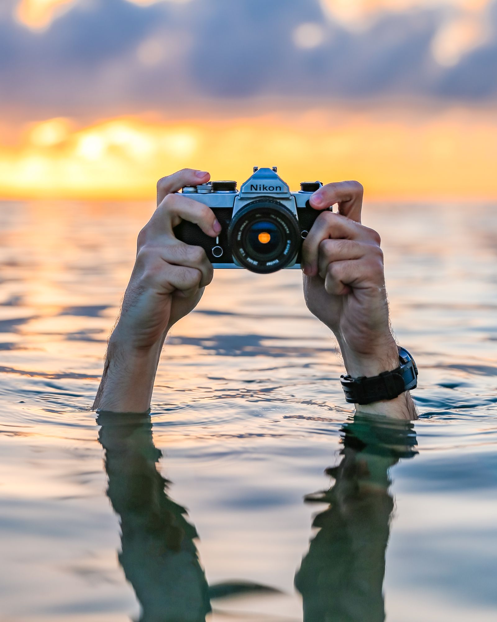 Two hands rising out of the water holding a film camera up to a sunset sky