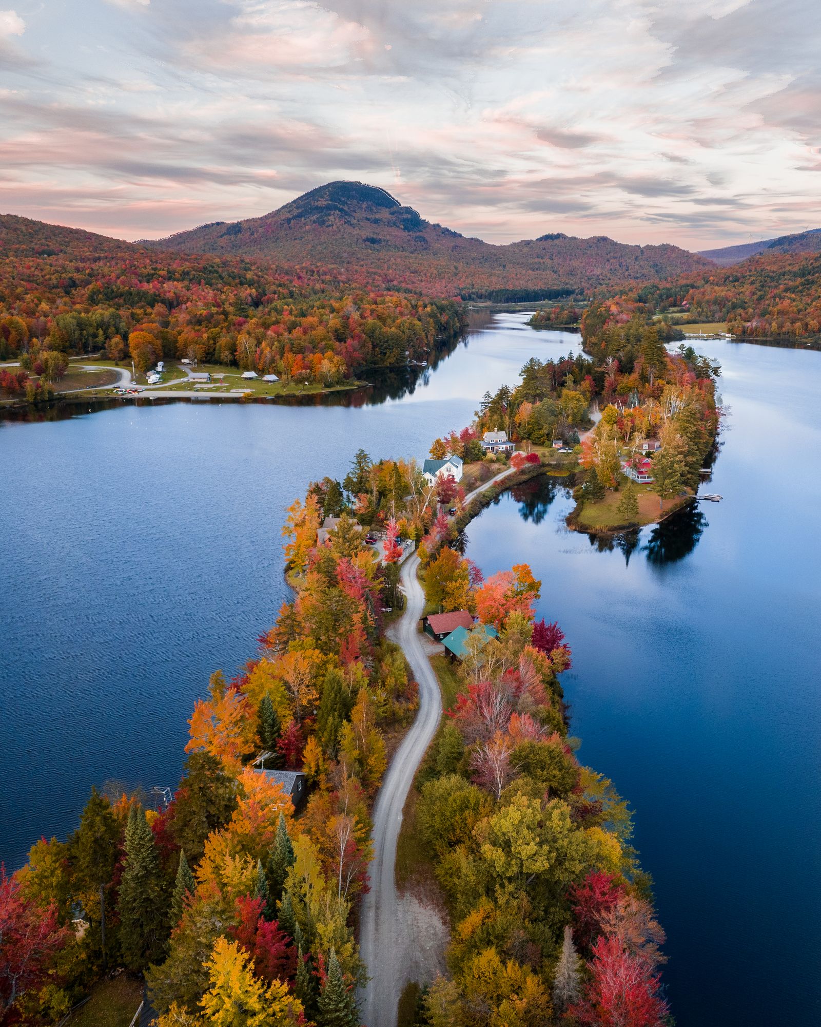 Aerial of a winding road along a wooded point between two lakes, the trees in full fall color under a mountain