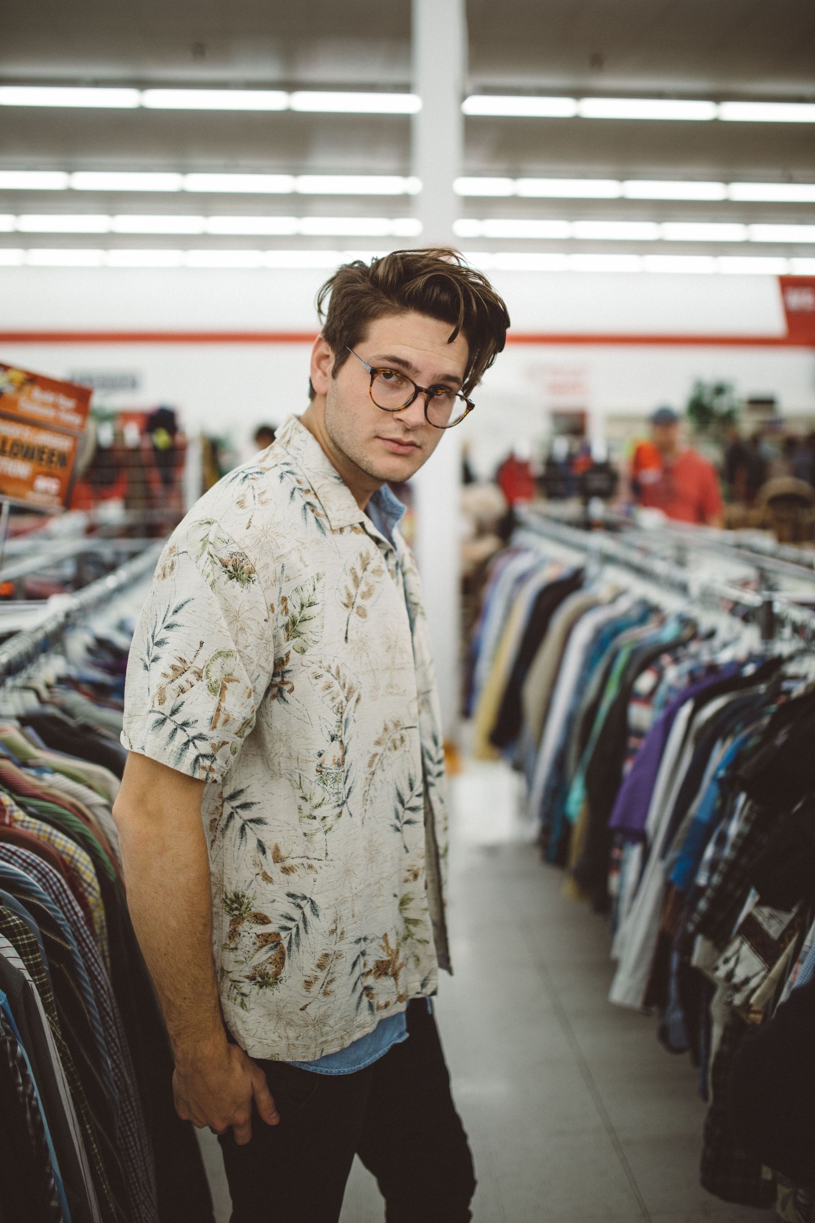 A young man in a printed shirt and glasses posing in a thrift store aisle