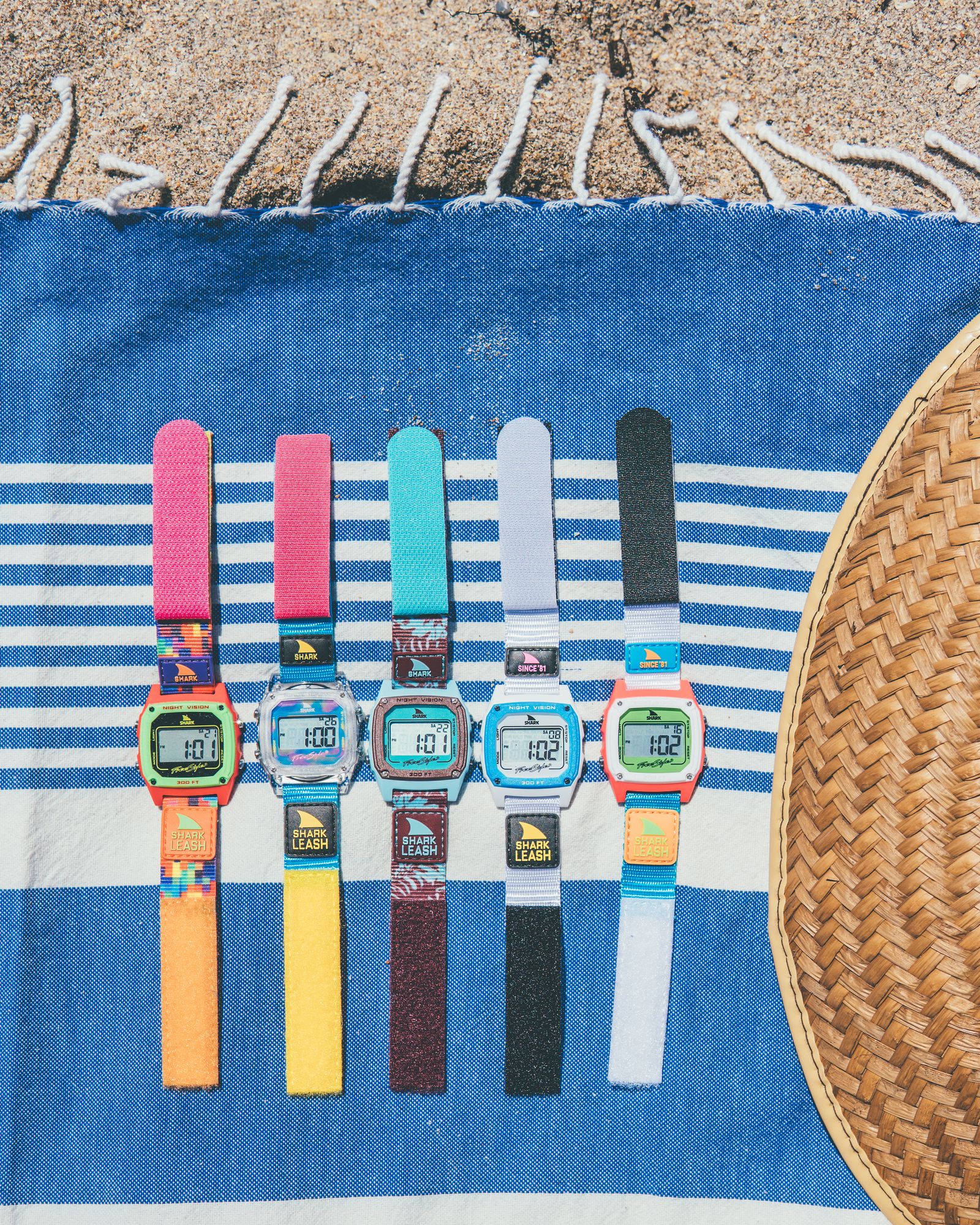 Five colorful Freestyle watches lined up on a striped beach towel beside a straw hat