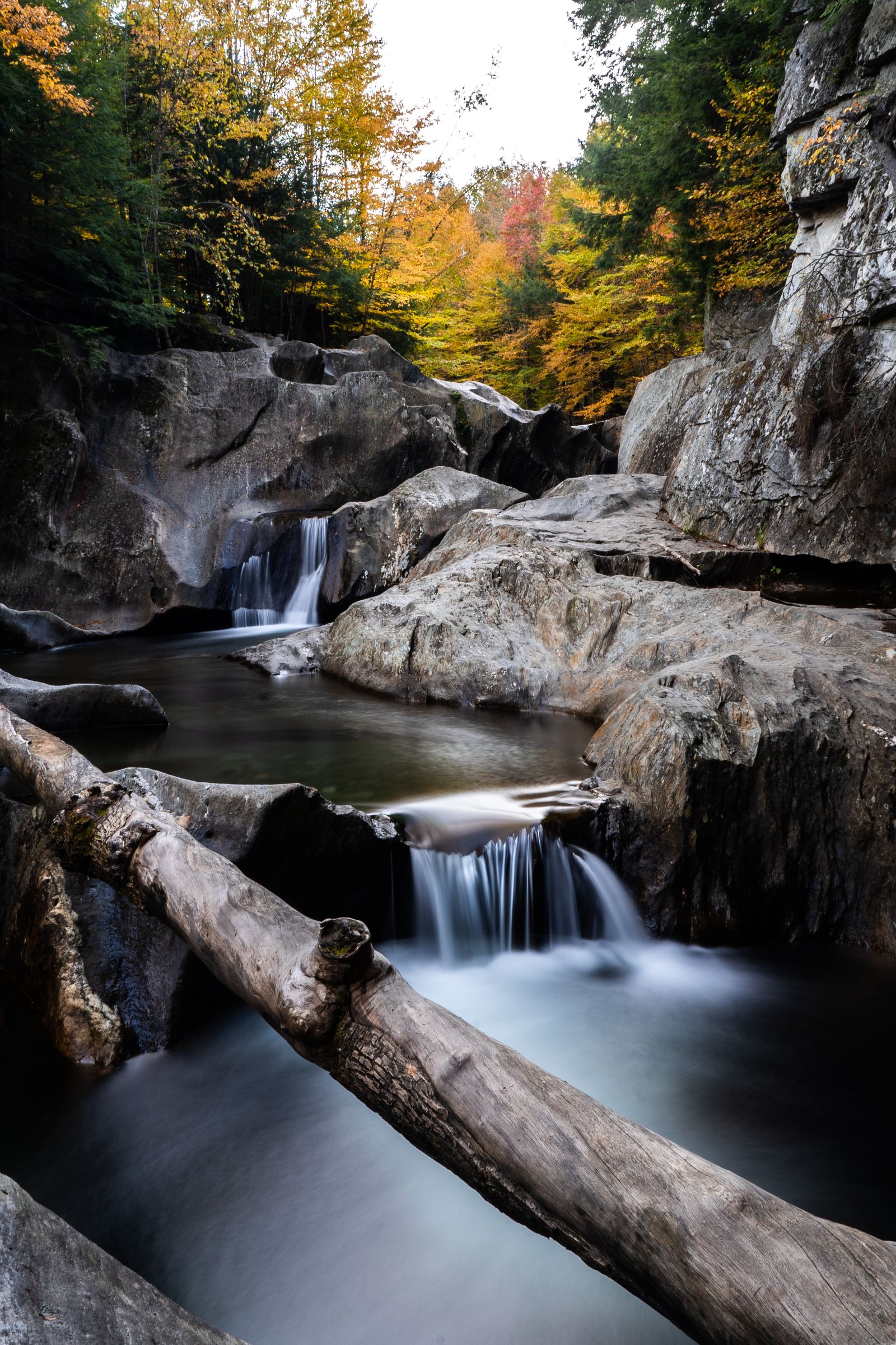 A long-exposure waterfall flowing over granite ledges past a fallen log, fall trees above